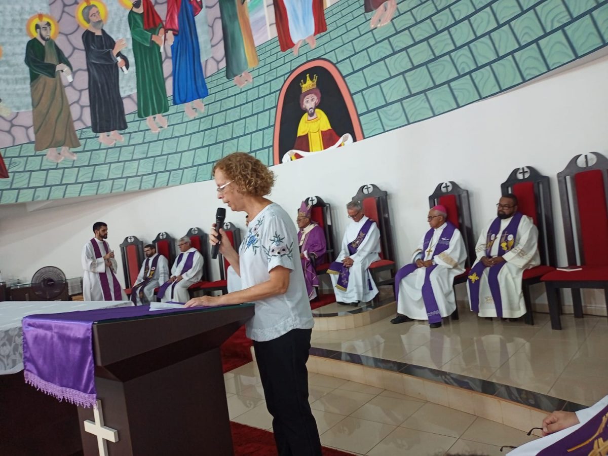 Foto durante uma celebração religiosa da Igreja Católica, na qual uma mulher está falando ao microfone e religiosos estão sentados com suas vestes brancas com detalhes roxos.